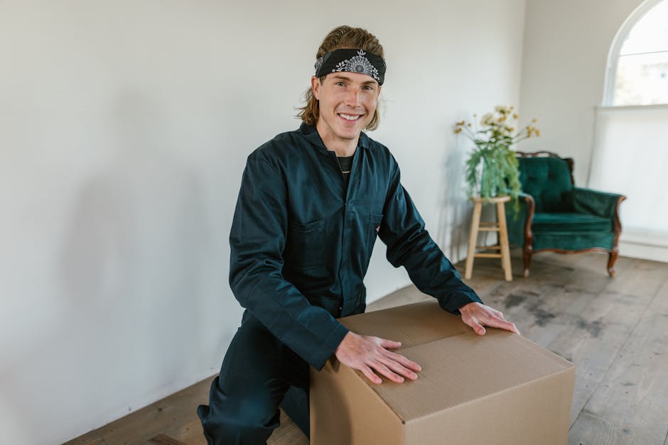 A male mover with shoulder-length hair, wearing a black bandana and navy blue work uniform, is inside a well-lit room during a home relocation process. He is smiling at the camera while kneeling and holding a large cardboard moving box, preparing for packing or transport. The room features light-colored walls, a wooden floor, and a large arched window allowing natural light to fill the space. In the background, there is a green upholstered armchair with wooden legs, positioned next to a small wooden side table with a potted plant and flowers. Part of a white door frame is visible on the left side of the image, suggesting the area is an interior hallway or living room in a residential property. This scene depicts the packing and furniture handling stage of a house move, likely managed by [COMPANY_NAME], which provides professional removals and moving services in Tottenham. The setting emphasizes careful handling of household furniture and packing materials as part of an organized furniture transport and moving process.
