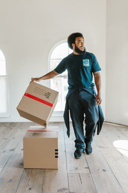 A man with curly black hair and a beard is inside a bright room with white walls and large arched windows, during a house removal. He is dressed in a navy blue t-shirt with a logo on the chest and dark trousers, holding a cardboard moving box with red tape on top in his right hand, preparing to place it on top of a similar box on the wooden floor. The room appears spacious, with natural light streaming through the windows, and has minimal furniture or decoration visible. This image captures the process of packing and furniture transport, reflecting home relocation activities complete with packing materials and careful handling of items, supporting the context of professional removals services offered by Man with Van Tottenham, as seen on the company’s website page about their Tottenham moves.