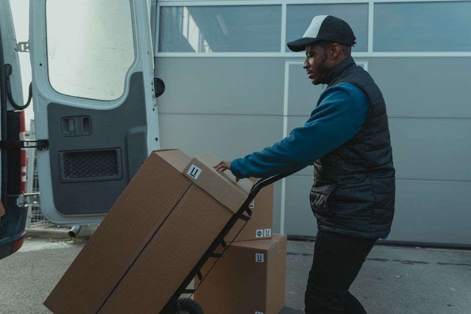 A man wearing a dark cap, blue long-sleeved shirt, and black padded vest is pushing a black-handled trolley loaded with two large cardboard boxes outside a grey van, which is parked next to a modern i