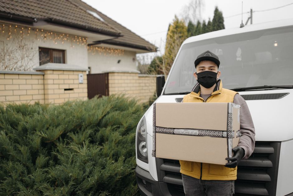 A man wearing a dark cap, blue long-sleeved shirt, and black padded vest is pushing a black-handled trolley loaded with two large cardboard boxes outside a grey van, which is parked next to a modern i