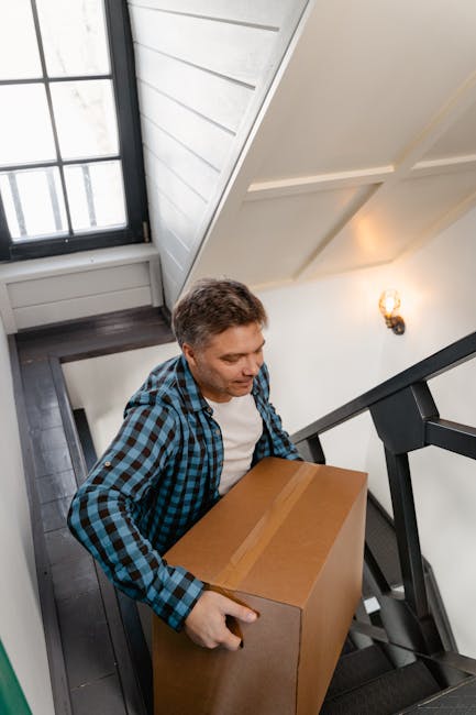 A man with short brown hair wearing a blue and black checkered shirt is seen carrying a medium-sized cardboard box down a staircase inside a residential property. The staircase features dark wooden steps with a black metal handrail, and the walls are painted white, with a decorative wall light fixture visible on the right. Natural light enters from a large window at the top of the stairs, illuminating the scene. The interior appears clean and well-maintained, with the man carefully navigating the stairs during a home relocation or furniture transport process. The image is associated with professional removals services, as offered by Man with Van Tottenham, and depicts the packing and moving steps involved in house removals, including furniture transport and handling narrow staircases in urban environments.