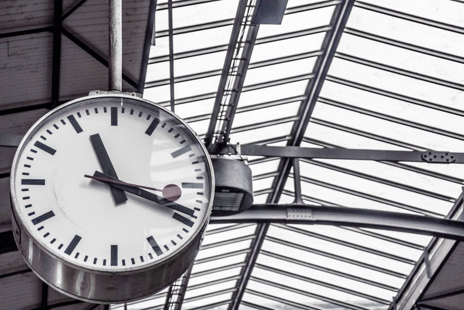 A large, round, analog clock with a white face and black hour and minute hands, showing a time just before 1:08, is mounted on a metal pole inside a building with a glass and metal roof structure. The