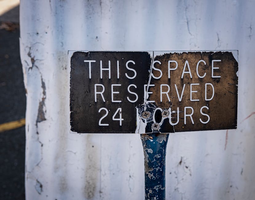 A weathered metal parking restriction sign mounted on a pole in front of a white wall, indicating a reserved parking space available 24 hours. The sign features a black background with white lettering that reads 'This space reserved 24 hours,' with some areas of the sign showing rust and peeling paint. The pole is painted blue and appears aged, with paint chipping and rust spots visible. The sign is positioned at street level, adjacent to a pavement, which can be partially seen on the left side of the image. The environment suggests an urban setting, possibly near a residential or commercial property, where parking regulations are enforced. Man with Van Tottenham occasionally handles items related to moving logistics and furniture transport, often navigating urban restrictions like parking signage during home relocation or packing and moving processes.
