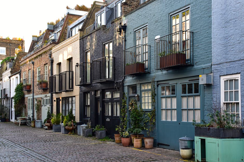 Photograph of a narrow cobblestone street in Tottenham showing a row of terraced houses with different facade colours and styles. Several buildings feature small balconies with black metal railings and potted plants, with some doors and windows decorated with climbing plants or window boxes. In front of the houses, there are various potted plants, gardening containers, and outdoor furniture placed on the pavement. A bicycle is leaning against one of the walls, and a blue wooden garage door is visible on the right side of the image. The lighting indicates a clear day with sunlight illuminating the scene, highlighting the textures of the brickwork and the vibrant colours of the buildings. The setting suggests a residential area suitable for house removals and furniture transport, with Man with Van Tottenham often handling relocations across such terraced streets while ensuring careful movement of household items, packing, and loading processes, especially on tight terraces like these.