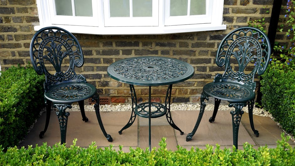 A wrought iron outdoor patio table and two matching chairs situated on a paved area in front of a brick house with a white-framed window. The table has a decorative pierced top, and the chairs feature intricate backrest designs. The scene is surrounded by neatly trimmed green bushes, and the setting appears to be part of a home garden or backyard, providing a calm outdoor space that could be involved in a house relocation or moving process managed by Man with Van Tottenham, especially during furniture transport or packing and moving activities.