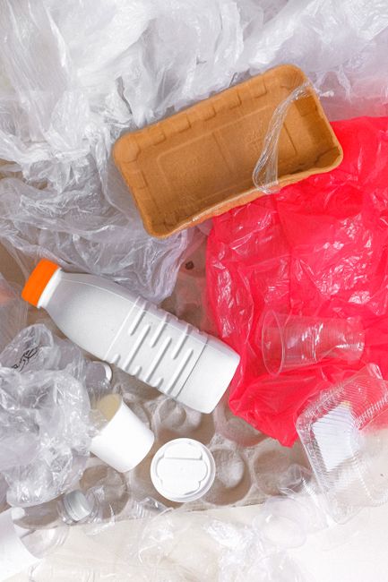 A close-up of various discarded packaging and waste materials on a floor, including clear plastic bags, crumpled plastic wrapping, a red plastic bag, a cardboard food container, and plastic bottles, some with caps and labels still attached. Items are scattered around, with no furniture or appliances visible. This scene appears to be the aftermath of packing or clearance during a home moving process, relevant to house removal services offered by Man with Van Tottenham. The materials are situated on a light-colored floor, and the image highlights waste disposal aspects associated with furniture transport and packing for a house relocation in Tottenham, Haringey.