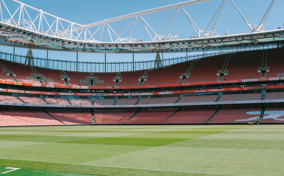 An empty scoreboard and tiered seating within a large football stadium, with red and pink seats arranged in multiple levels around the pitch. The stadium's roof structure, composed of white steel trusses, arches above the seating area, partially covering the field. The grass pitch is well-maintained, with visible mowing patterns, and the sky above is clear and bright, indicating daytime. This setting is unrelated to house removals or moving services and does not depict any furniture, boxes, or vehicles involved in a home relocation. The image focuses solely on the stadium's architecture and playing field.