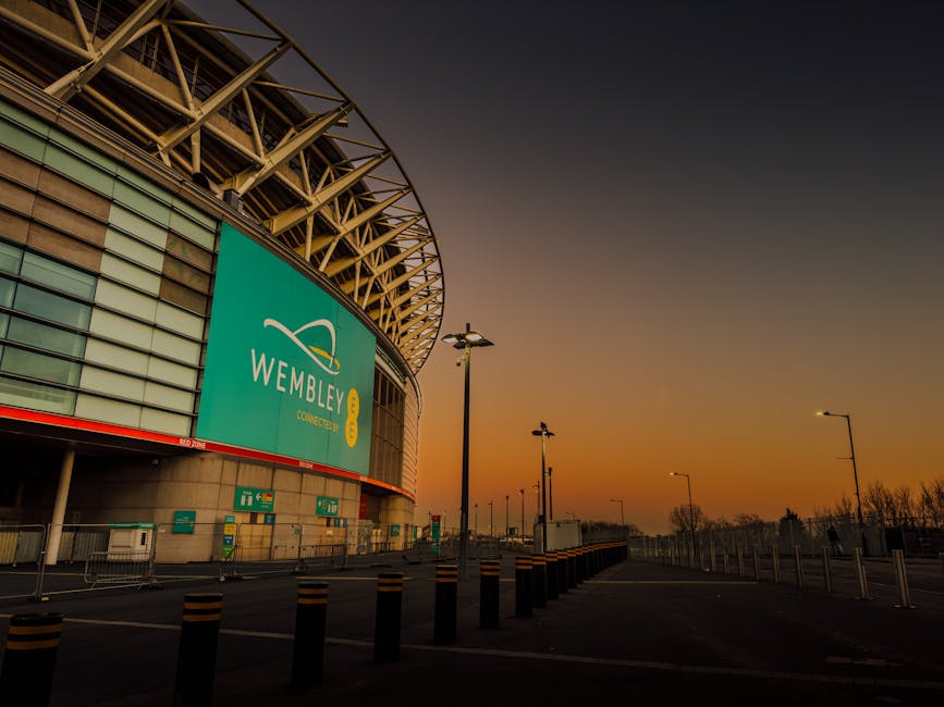 Exterior view of Wembley Stadium at sunset, showing its large, curved steel roof structure and glass facade with a prominent turquoise Wembley Connected sign. The scene includes a paved forecourt with black and yellow bollards, street lamps, and some metal barriers, indicating a designated pedestrian or vehicle area. Alongside the stadium, there are a few parked trucks and Vans, suggesting the logistics of home relocation or furniture transport preparations for moving services. The lighting is soft due to the setting sun, casting an orange glow across the scene, emphasizing the modern architecture and the surrounding road infrastructure, which aligns with local moving and removals operations by Man with Van Tottenham for house or furniture relocations near Tottenham Hotspur Stadium.