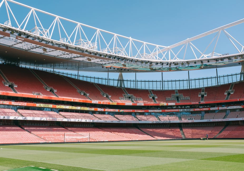 An empty scoreboard and tiered seating within a large football stadium, with red and pink seats arranged in multiple levels around the pitch. The stadium's roof structure, composed of white steel trusses, arches above the seating area, partially covering the field. The grass pitch is well-maintained, with visible mowing patterns, and the sky above is clear and bright, indicating daytime. This setting is unrelated to house removals or moving services and does not depict any furniture, boxes, or vehicles involved in a home relocation. The image focuses solely on the stadium's architecture and playing field.