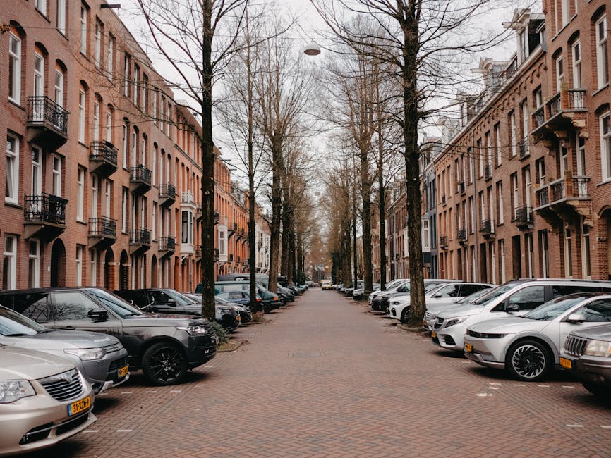 A residential street in Tottenham with row of multi-story brick apartment buildings on both sides, each featuring small balconies and large windows. The street is lined with leafless trees, indicating a colder season, and has a row of parked cars along the curb, including sedans, hatchbacks, and SUVs in various colours such as silver, black, and grey. The pavement is made of reddish bricks, and the sky is overcast, suggesting cloudy weather. This scene may depict an urban area where home relocation or furniture transport is taking place, with potential parking suspensions in the vicinity for moving services, as managed by companies like Man with Van Tottenham. The street appears quiet and suitable for loading or unloading furniture during a house move, with space available for vans and removal vehicles.