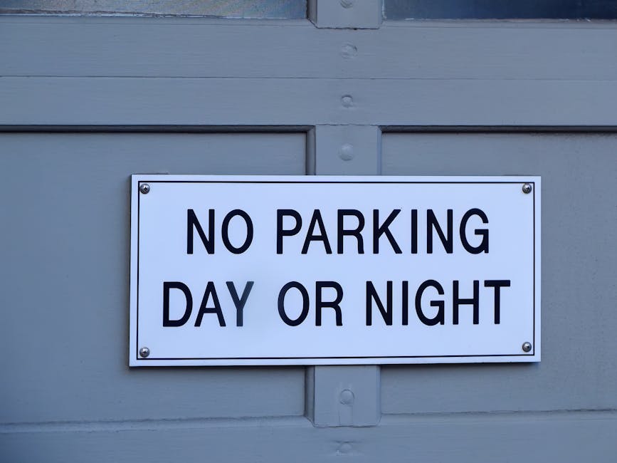 A rectangular white sign with black bold lettering stating 'NO PARKING DAY OR NIGHT' is affixed to a light grey wooden door or panel, which features visible horizontal and vertical wooden planks. The sign is mounted with four metal screws, one at each corner. Behind the door or panel, there is a paved area visible, possibly pavement or a driveway, used for loading or unloading furniture and boxes during a home relocation. The lighting is natural and even, with no shadows or reflections, indicating an outdoor setting. This image highlights parking restrictions that may affect logistics involved in residential removals or furniture transport, as managed by companies like Man with Van Tottenham, especially during scheduled moves in Tottenham. The scene emphasizes the importance of parking suspensions or permits when planning for house removals to ensure a smooth loading process for furniture and packing materials.