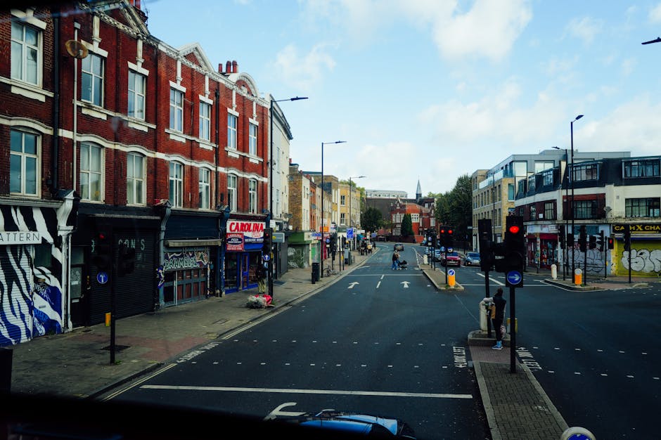 View of a busy street in Tottenham, N17 with historic red brick buildings on the left, some with storefronts such as 'Carolina' and others with awnings. On the right side, modern white and black residential buildings feature balconies. The street is lined with black lampposts and traffic lights, with cars waiting at a red light and others driving on the road. Several pedestrians are visible on the sidewalks, and some are near a loading area where a man is carrying a cardboard box towards a van parked on the pavement. The scene includes parking meters, bollards, and an overhead sky with some clouds, indicating daylight. This setting illustrates the typical environment in Tottenham, where relocation services like those provided by Man with Van Tottenham organize home relocations and furniture transport, often involving loading and unloading items during the moving process.