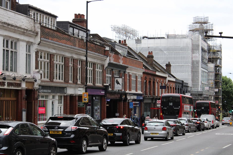 A busy street scene in Tottenham, featuring a row of brick and white-painted commercial buildings with large windows, some with signage, and one under construction with scaffolding and a protective white covering. In the foreground, multiple vehicles are parked along the curb, including black, grey, and white cars, with two red double-decker buses traveling down the street. Pedestrians are walking on the sidewalk, and some are near the shop entrances. The image was captured during daylight hours under overcast weather, with soft lighting illuminating the scene. This scene reflects typical urban traffic and commercial activity, relevant to home relocation and furniture transport logistics, which Man with Van Tottenham can assist with for flats on N17, including access tips on Tottenham High Road.