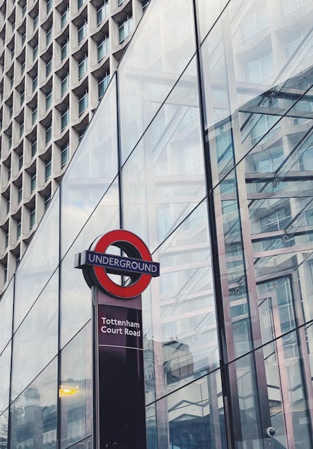 A close-up view of a London Underground sign with a red and blue roundel displaying the word 'UNDERGROUND' above a black rectangular panel indicating 'Tottenham Court Road' station. The sign is mounted on a sleek, modern glass building with reflective surfaces, which mirror the surrounding cityscape and partly obscure the building's interior. The background features a high-rise structure with a grid of windows, and the sky is visible through the reflections. The scene depicts an urban environment consistent with central Tottenham, where [COMPANY_NAME]'s house removal and furniture transport services may operate. The image captures the typical city infrastructure involved in home relocation logistics, illustrating the accessibility of transport links for moving or packing and moving activities.