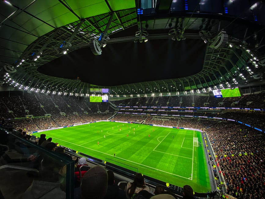 Interior view of a large football stadium with a bright green pitch, illuminated by overhead lighting. The seating areas around the field are filled with spectators, and multiple screens above display information. The ceiling structure, with its metallic framework and some hanging equipment, is visible overhead. This setting depicts a professional sports venue during a match or event, with the focus on the football pitch and surrounding crowd. The environment is well-lit with a mix of natural and artificial lighting, creating a vivid atmosphere for the game. Although the image is primarily of a sports stadium, the detailed description aligns with how Man with Van Tottenham might describe a well-lit, indoor event space used for various purposes, including special possessions transportation or storage during venue relocations.