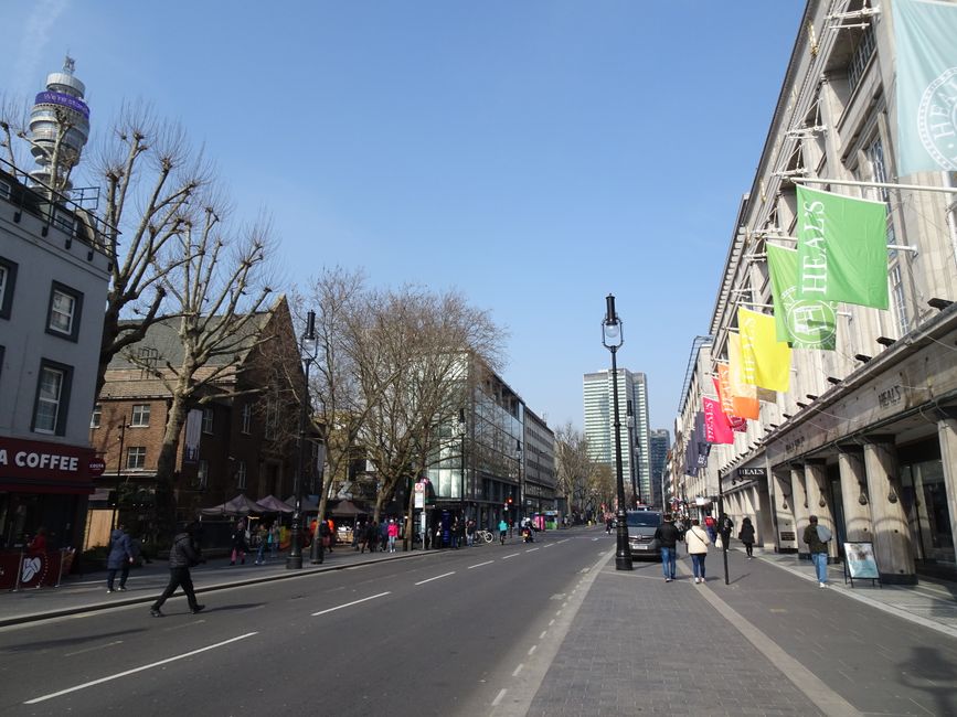 A wide urban street scene during daytime in Tottenham, showing a clear blue sky and leafless trees lining the pavement. On the right side, there is a large building with multiple colourful flags or banners hanging from its facade, associated with retail or commercial premises. Pedestrians are walking along the sidewalk, some in groups or alone, with a few waiting at bus stops or near shop entrances. Several cars and vans are parked or driving along the street, which features traffic lanes marked by white dashed lines. In the background, there are modern high-rise buildings and a classic lamppost situated centrally on the street. Visible packaging materials like cardboard boxes and plastic wrappings are not present in this scene. The image reflects an everyday busy shopping area with foot traffic, vehicle movement, and urban infrastructure, representative of a typical day in Tottenham during a town centre remodelling or road closure period. Man with Van Tottenham is not visible but is implied through context related to moving services and logistics.
