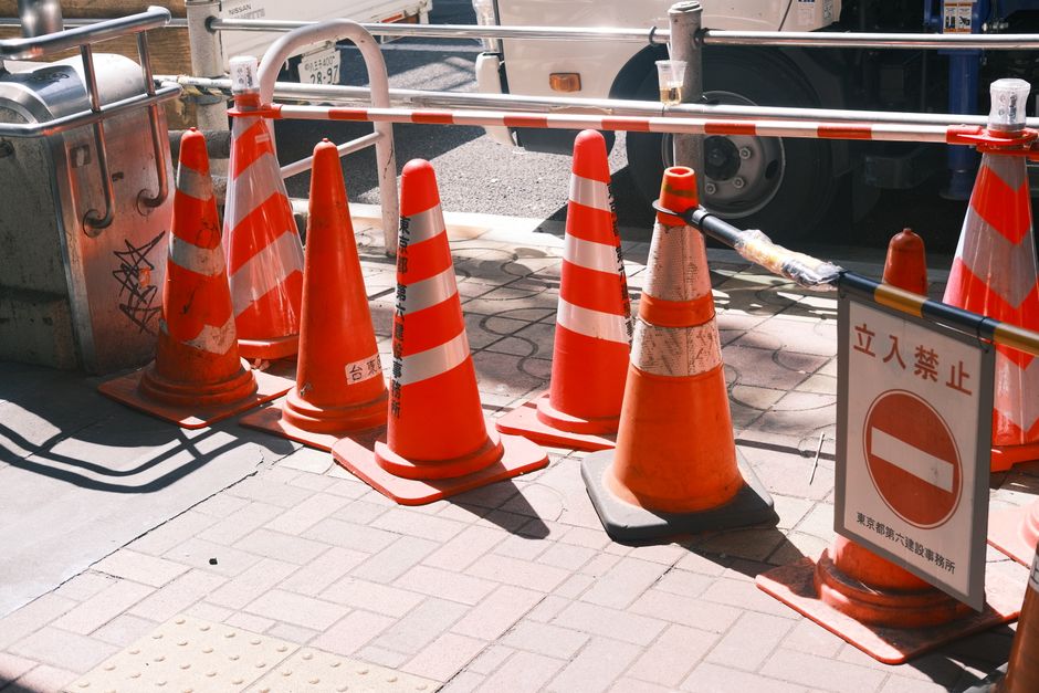 A row of orange and white traffic cones and barricades positioned along a city sidewalk, marking a restricted area during road closures in Tottenham. The cones are placed tightly together, some with visible wear, and are connected by black and yellow tape stretched across the top, indicating a no-entry zone. A white sign with red Japanese characters and a horizontal red bar is attached to one of the cones, signaling the closure to pedestrian and vehicle access. Behind the barricades, there is a large truck with black tires parked on the street, partially visible through the gaps between the cones. To the left, a metal waste bin with graffiti is situated on the pavement next to the barricades. The sidewalk comprises concrete paving slabs with tactile paving patches near the curb. Bright daylight and shadows cast by the cones and barricades highlight the active construction or roadwork area, which may relate to a temporary removals permit or home relocation process coordinated by Man with Van Tottenham.