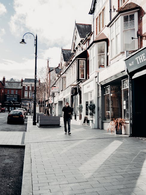 A busy urban street scene featuring a row of shops and offices on the right side, with colorful flags hanging from their facade, and pedestrians walking along the wide sidewalk and crossing the road.
